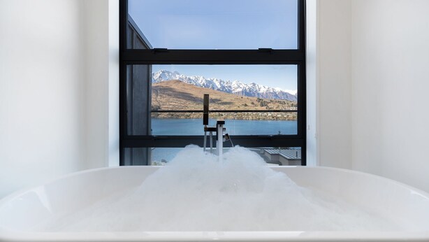 A bathroom with a bathtub filled with water, a window, and a view of mountains and a lake. - St Matthews Views - Cecil (Queenstown)