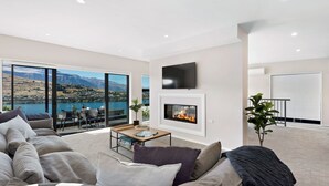 A modern living room with a fireplace, a flat-screen TV, and a view of a lake and mountains. - St Matthews Views - Cecil (Queenstown)