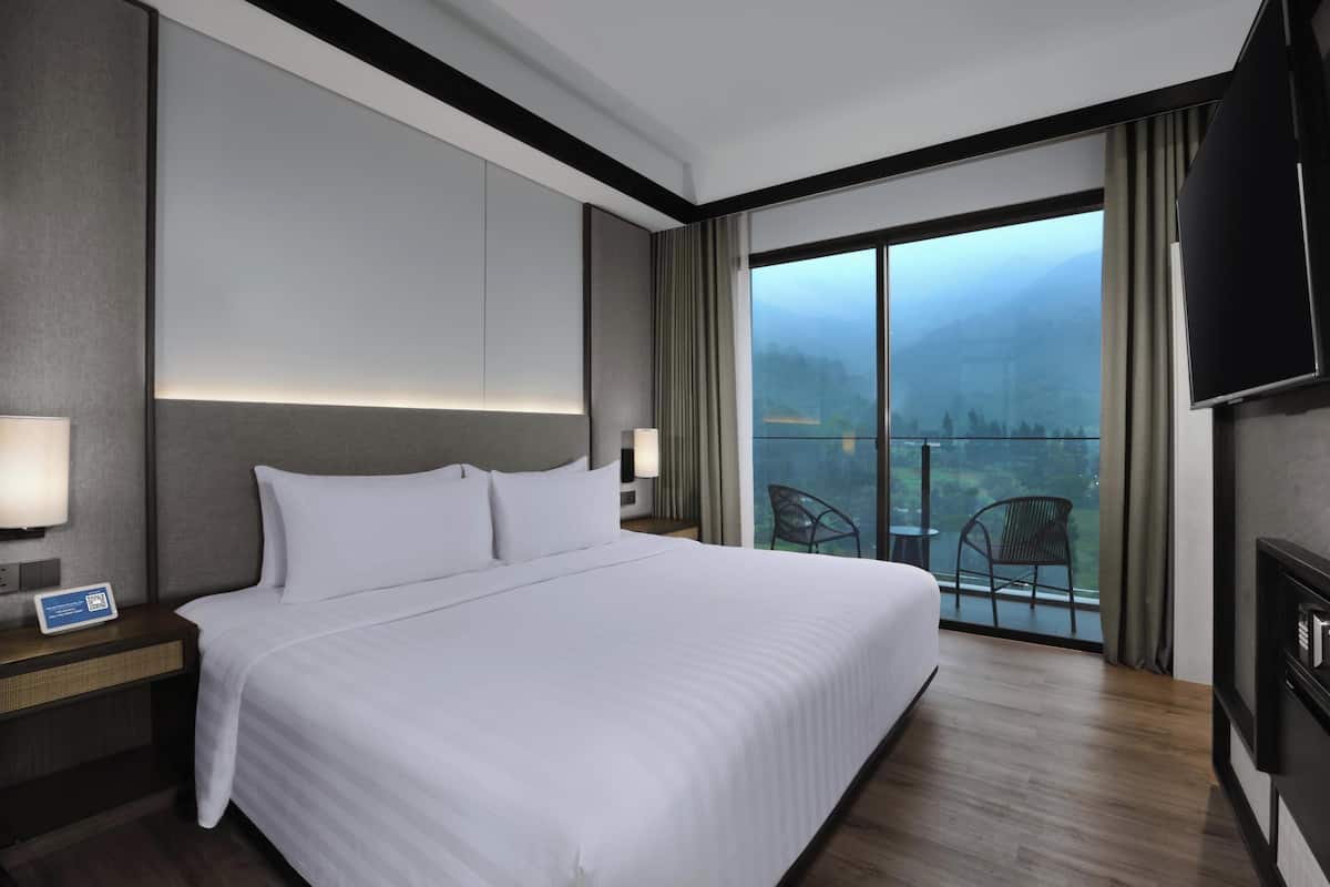 A hotel room with a large bed, a flat-screen TV, a balcony with a view of mountains, and a desk with a lamp.