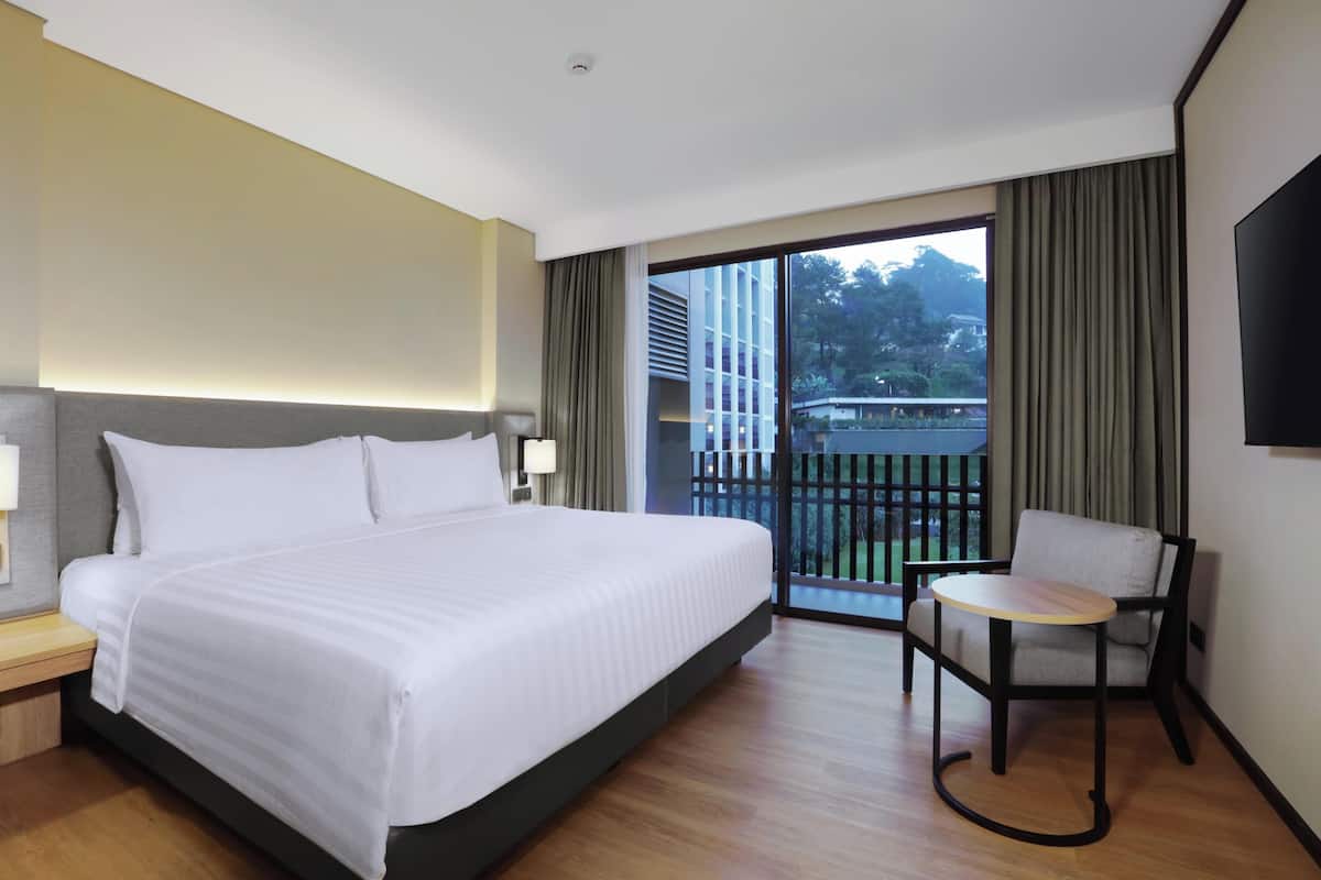 A hotel room with a large bed, a small table, a chair, and a balcony with a view of buildings and trees.