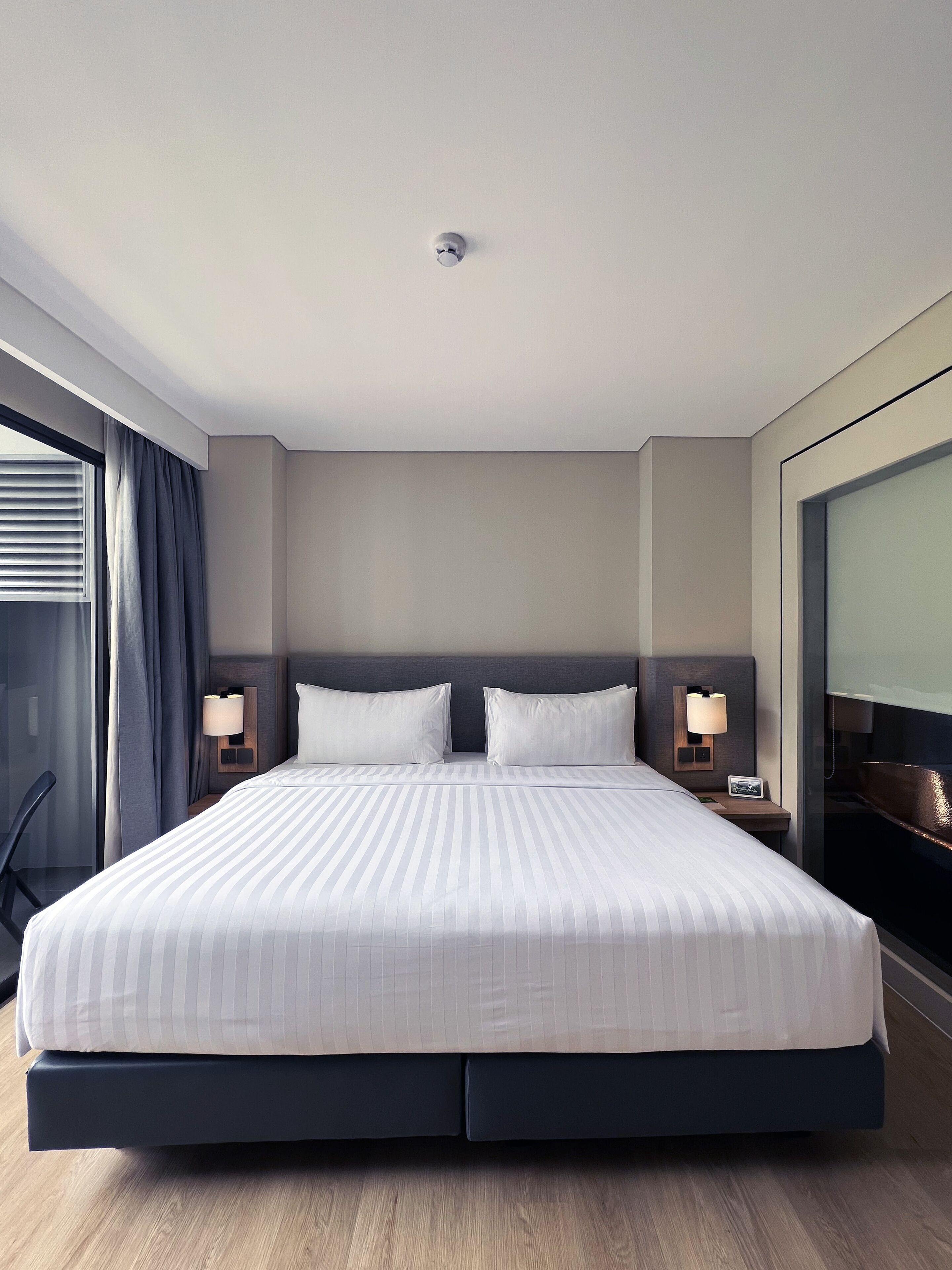 Una habitación de hotel moderna con una cama grande, dos mesitas de noche con lámparas, un ventanal con cortinas y vistas a la ciudad.