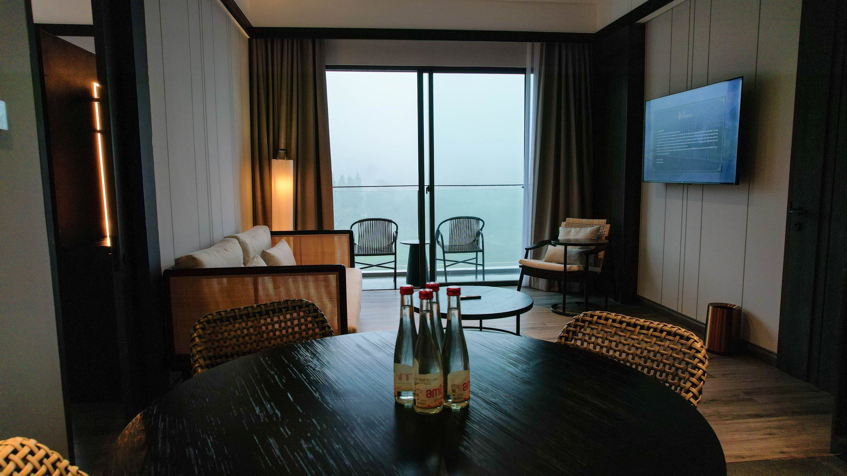 A hotel room with a balcony, a sofa, two chairs, a dining table with bottles, and a TV mounted on the wall.