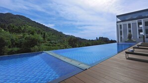 Una piscina infinita con terraza de madera y sillones de descanso, con vistas a una colina verde exuberante.