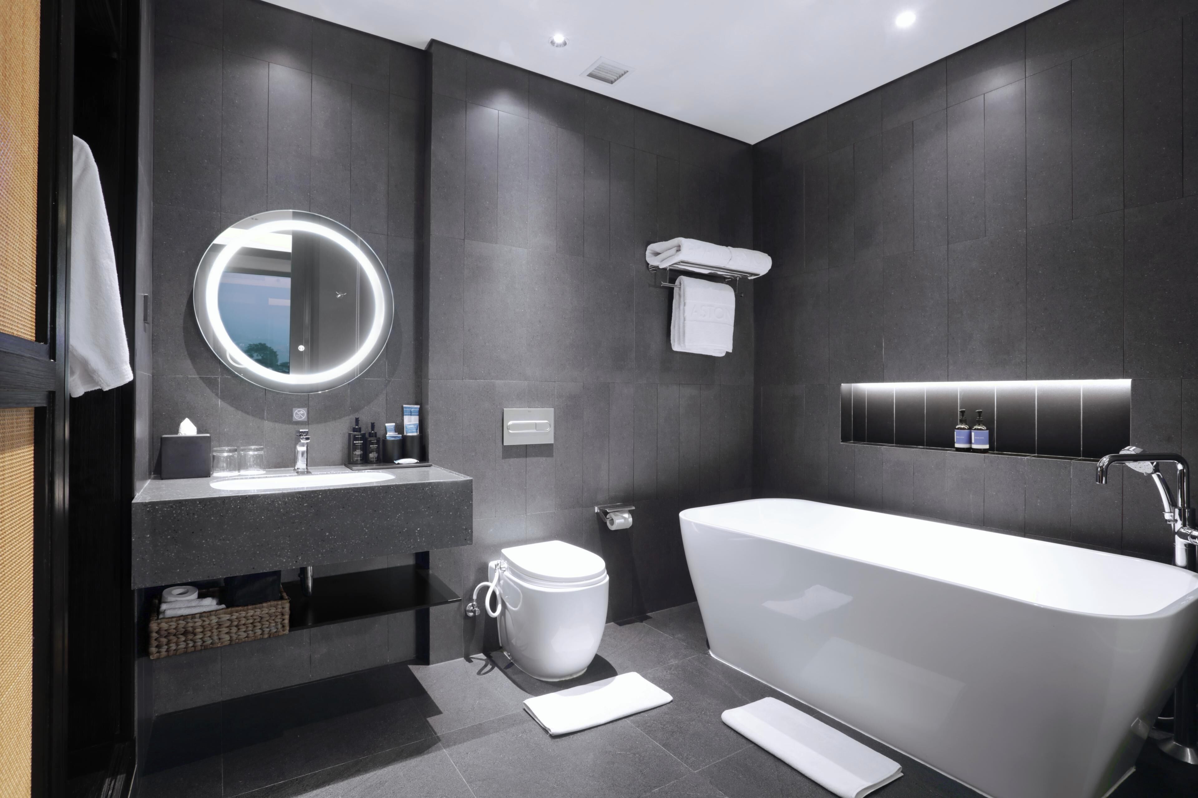 Ein modernes Badezimmer mit einer freistehenden Badewanne, einem runden Spiegel mit Beleuchtung, einer Toilette und einem Handtuchhalter.