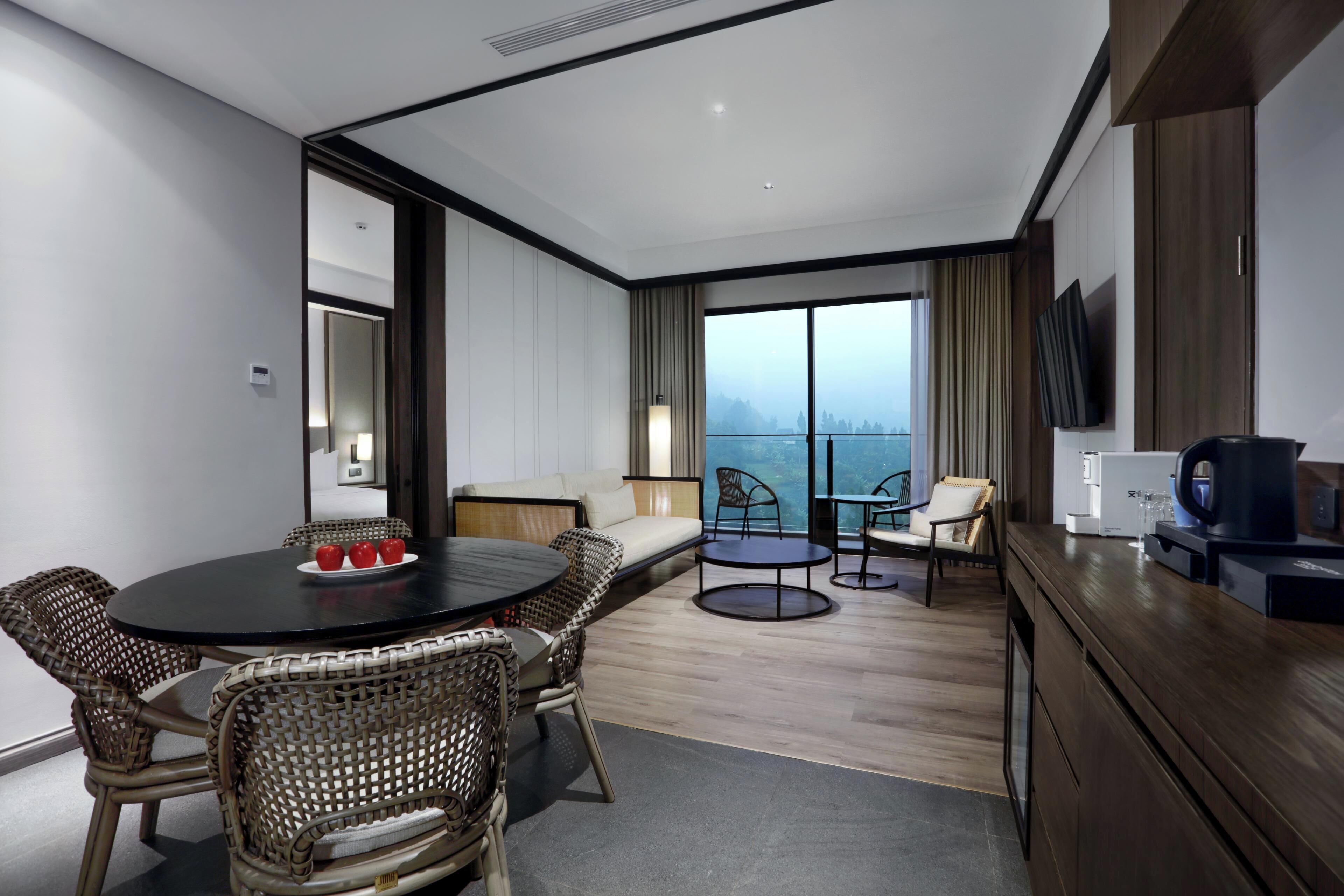 A modern hotel room with a dining area, a sofa, a coffee machine, and a view of the city.
