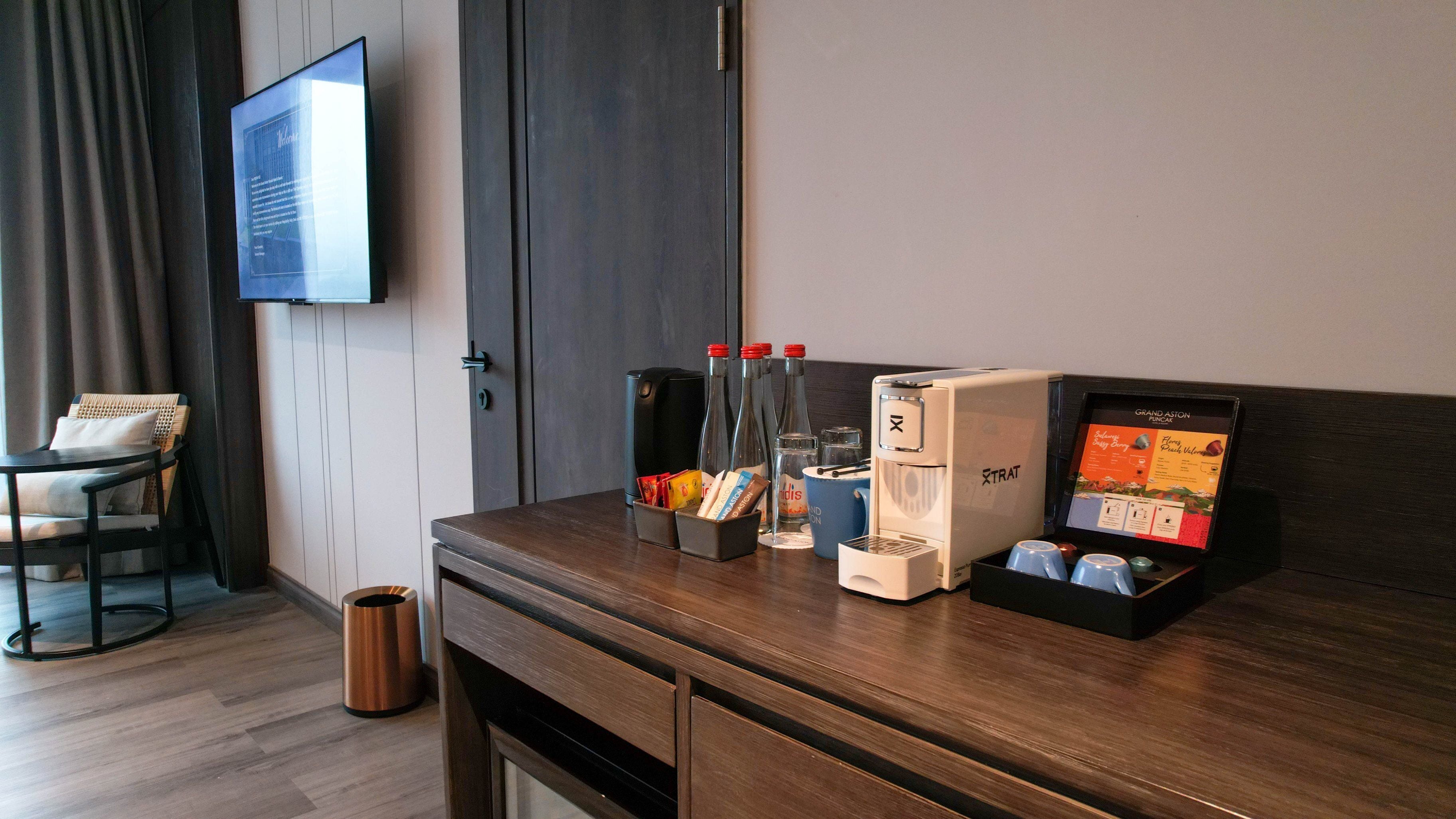 A hotel room with a wooden desk, a television mounted on the wall, a minibar, and a coffee machine.
