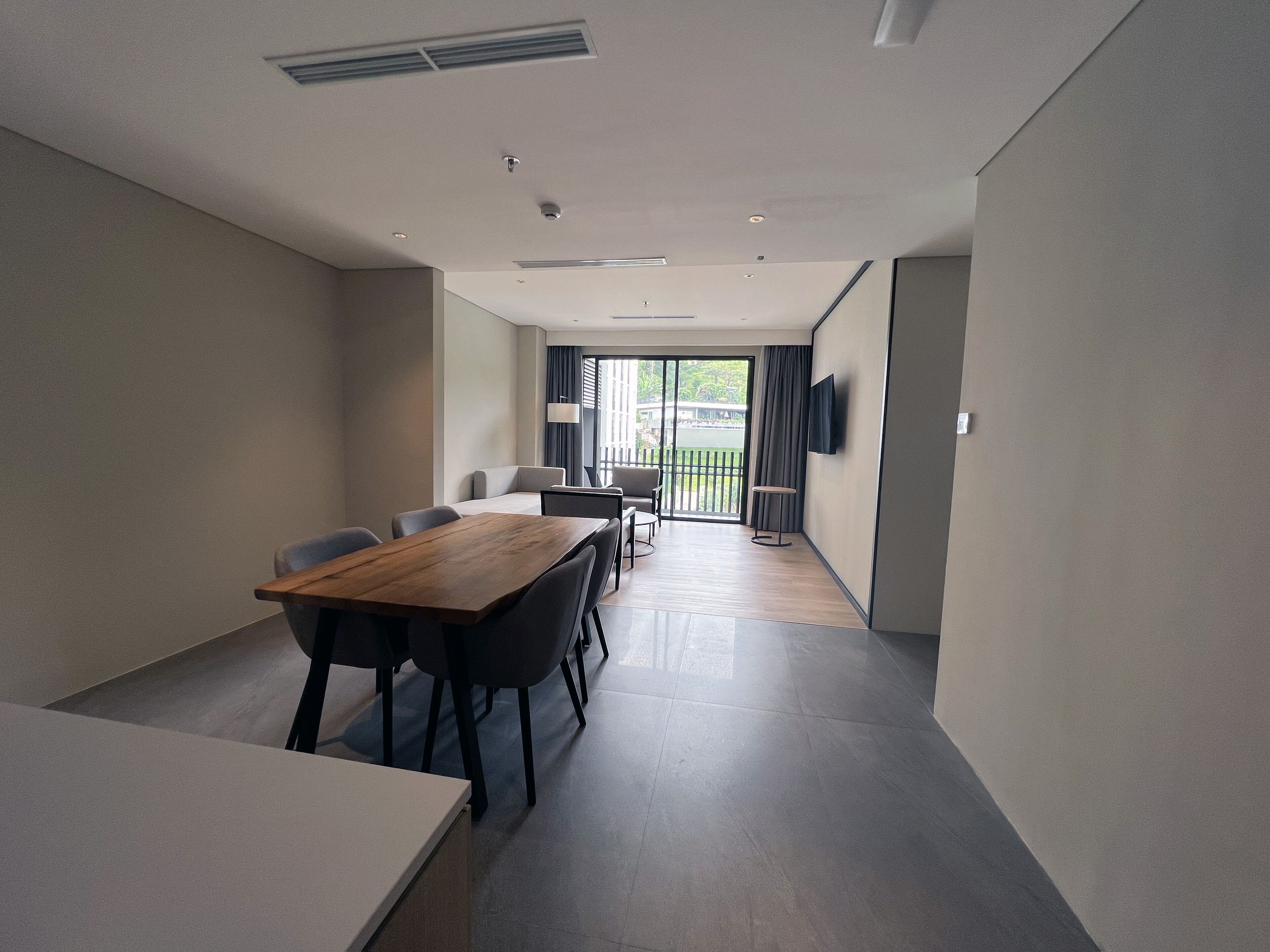 A modern room with a wooden dining table, black chairs, a flat-screen TV mounted on the wall, and a sliding glass door leading to a balcony.