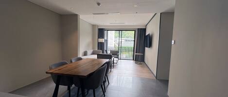 A modern room with a wooden dining table, black chairs, a flat-screen TV mounted on the wall, and a sliding glass door leading to a balcony.
