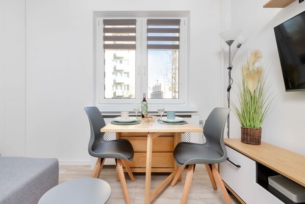 A modern dining area with a wooden table, two chairs, a window with blinds, a TV on the wall, and a plant on the side table.