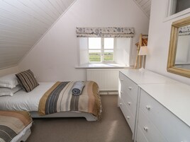 A bedroom with a sloped ceiling, a window with a floral valance, a bed with striped bedding, a bedside table with a lamp, and a dresser.