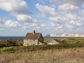 A stone house with a chimney, surrounded by grassy fields and a cliff in the background.