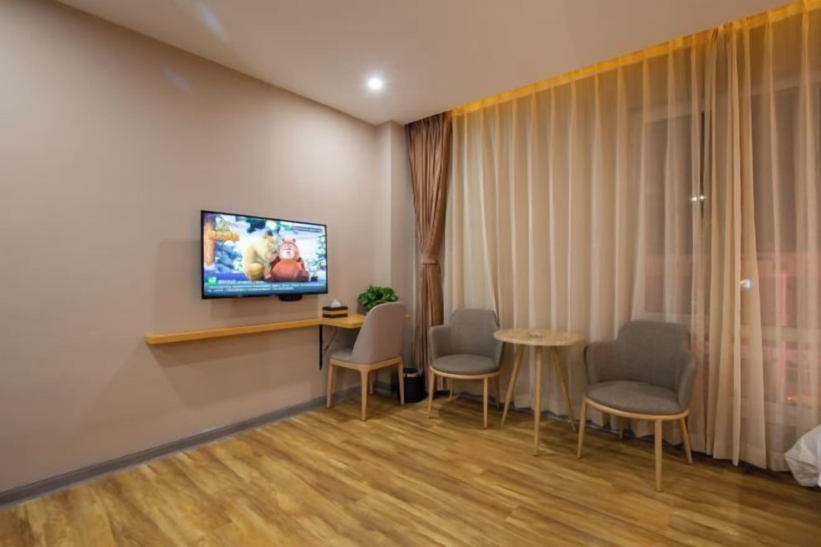 Een hotelkamer met een televisie aan de muur, twee stoelen, een klein tafeltje en een groot raam met gordijnen.