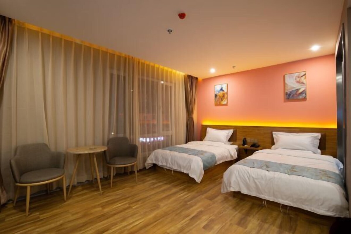 Una habitación de hotel con dos camas, una mesita pequeña, dos sillas y un ventanal grande con cortinas.