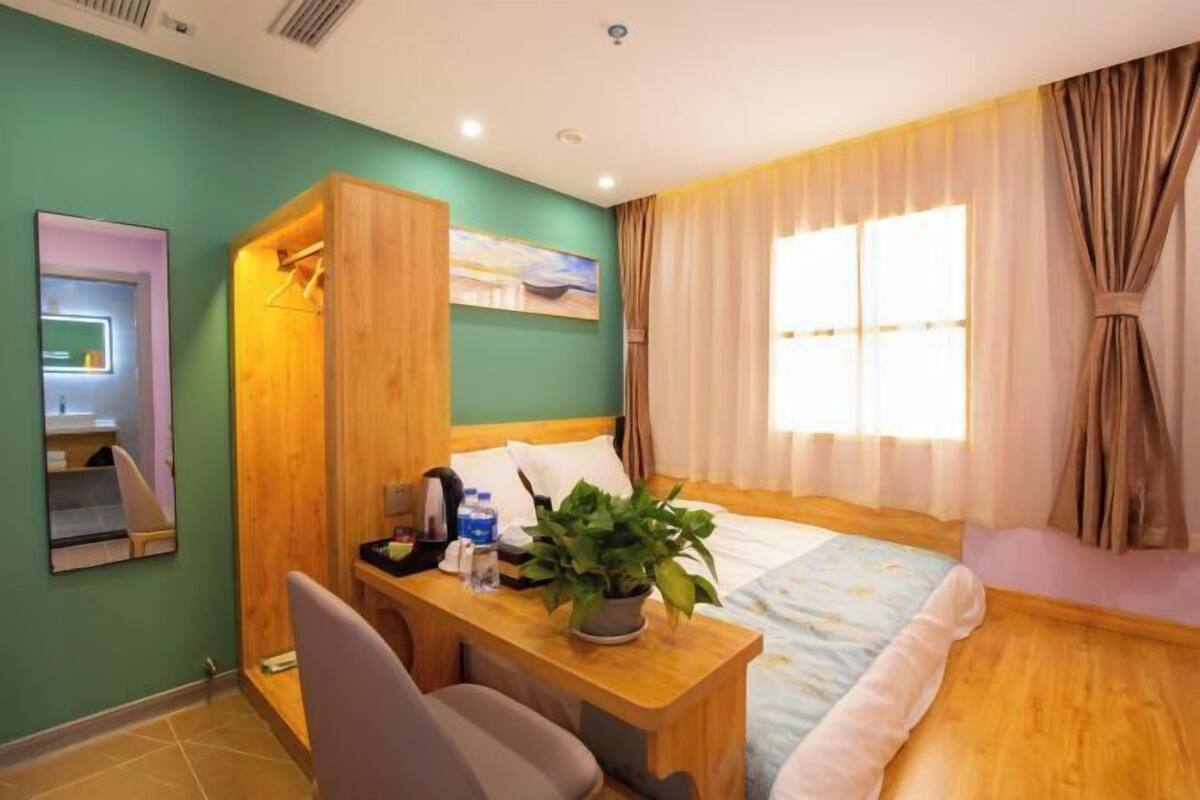 Quarto de hotel com escrivaninha de madeira, cama com colcha florida, planta em vaso e um quadro na parede.