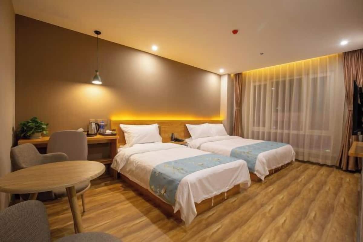 Quarto de hotel com duas camas, piso de madeira, uma mesa redonda, uma escrivaninha com abajur e uma janela com cortinas.