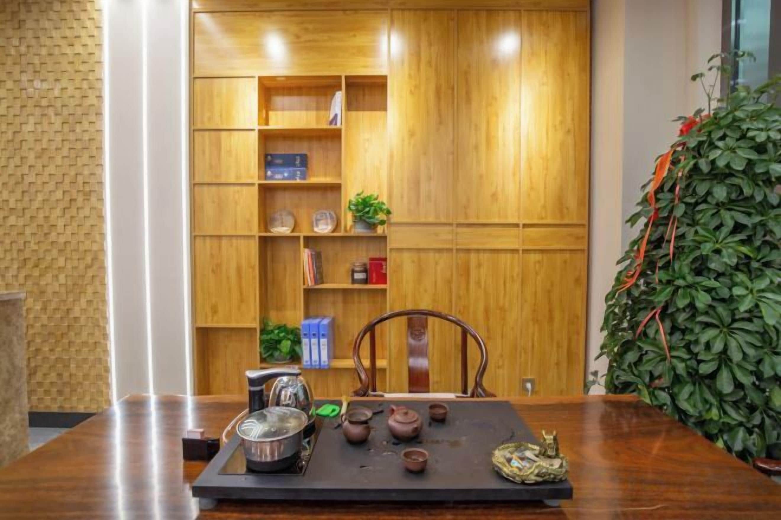 Una mesa de madera con pots de té y un juego de tetera, una silla y una estantería con varios objetos.