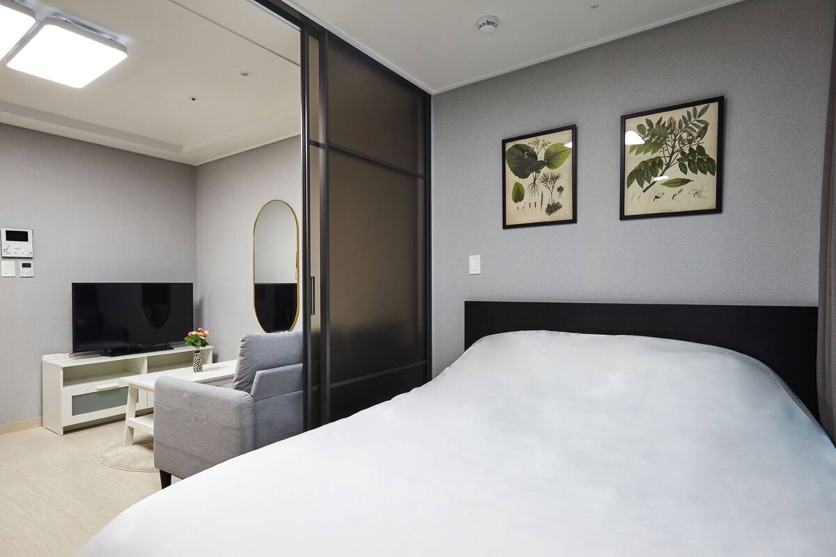 Een moderne hotelkamer met een groot bed, een flatscreen televisie, een bureau met stoel, en twee ingelijste botanische prints aan de wand.