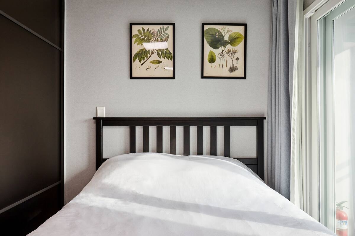 Een bed met een wit matras en een zwart hoofdbord in een kamer met een grijze muur. Twee ingelijste botanische prints hangen aan de muur.