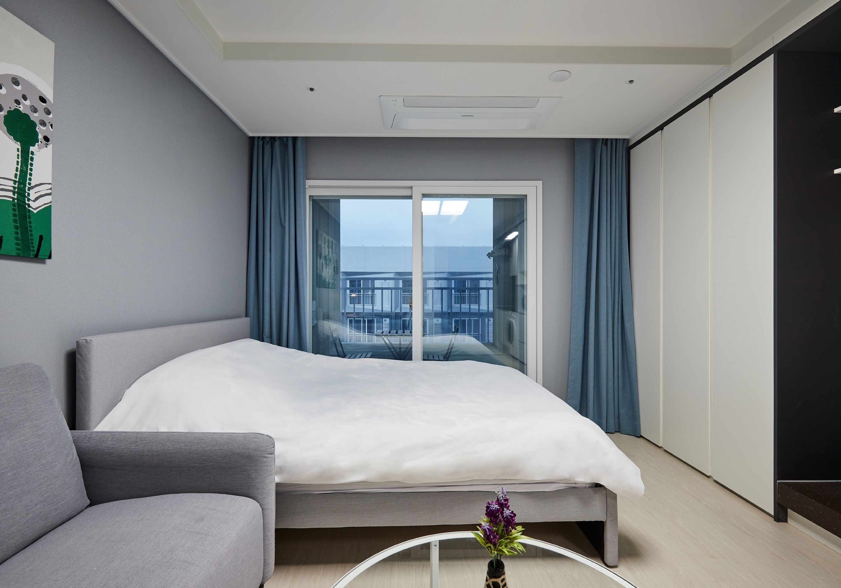 Una habitación de hotel moderna con cama, sofá, una pequeña mesa con un florero, un ventanal con cortinas y vistas a la ciudad.