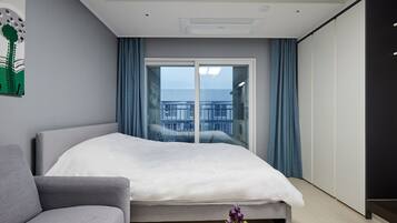 Una habitación de hotel moderna con cama, sofá, una pequeña mesa con un florero, un ventanal con cortinas y vistas a la ciudad.