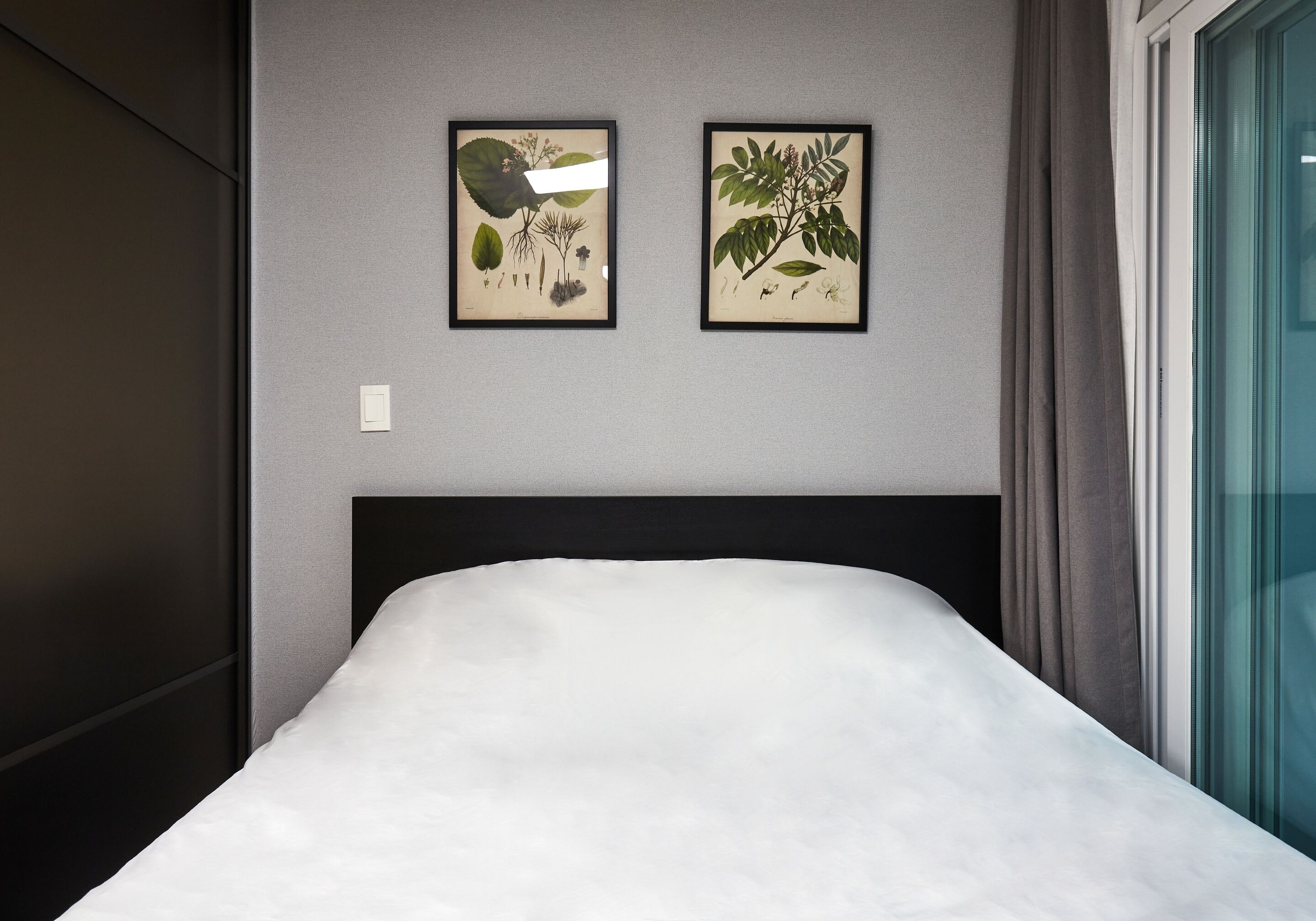Una cama bien hecha con sábana blanca en una habitación moderna con paredes grises y dos impresiones botánicas enmarcadas encima.