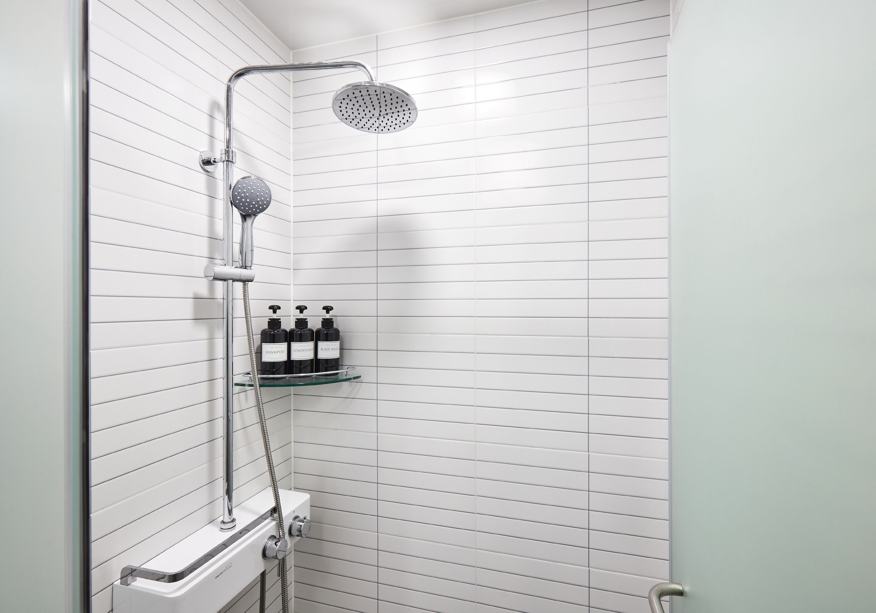 A modern shower with a handheld showerhead and a wall-mounted rain showerhead. There are two bottles of shampoo and conditioner on a glass shelf.