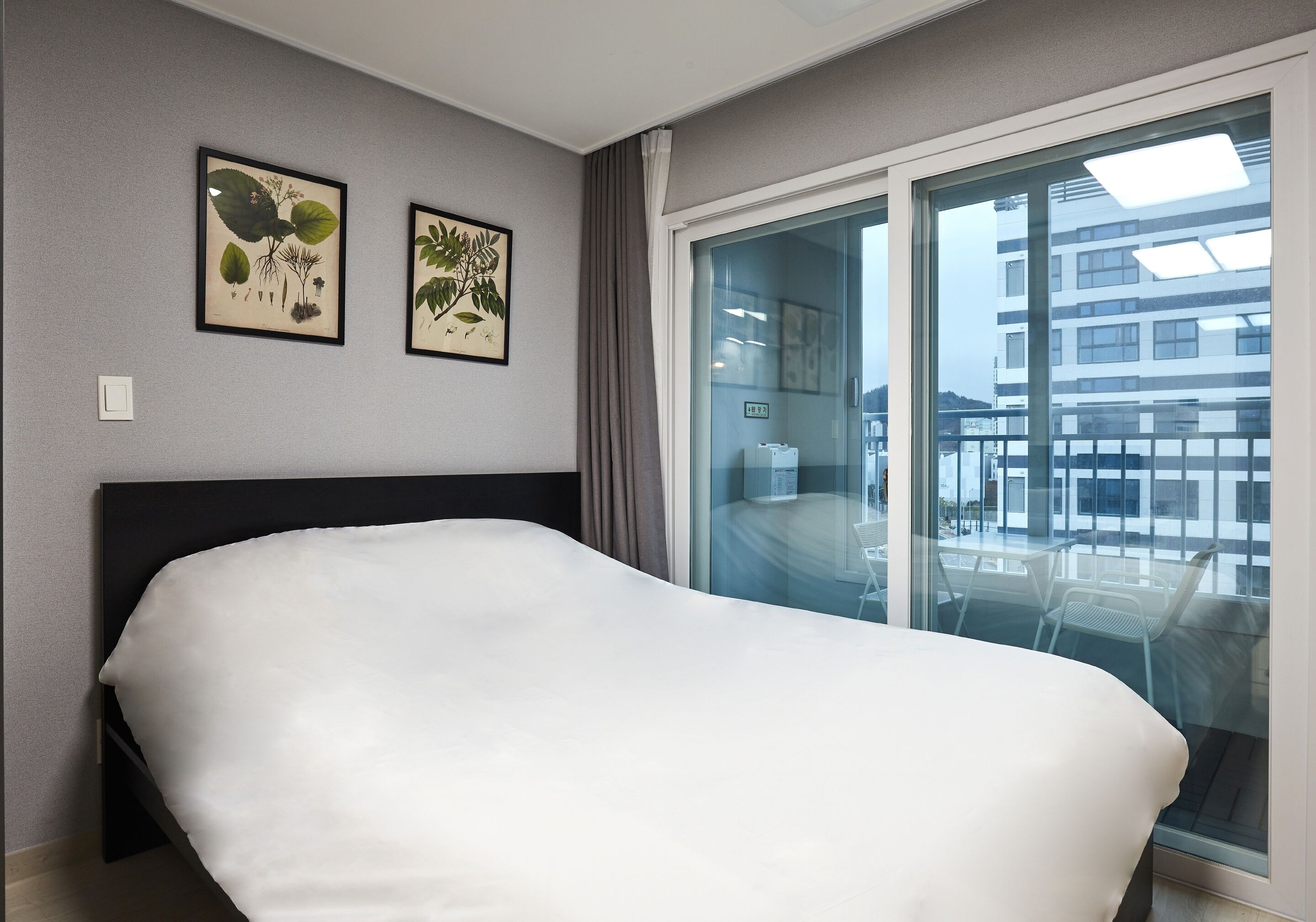 Quarto de hotel com uma cama, duas gravuras botânicas emolduradas na parede e uma varanda com vista para edifícios.