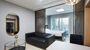 A modern hotel room with a glass table, a black leather sofa, a large mirror, and a view of the city through a large window.