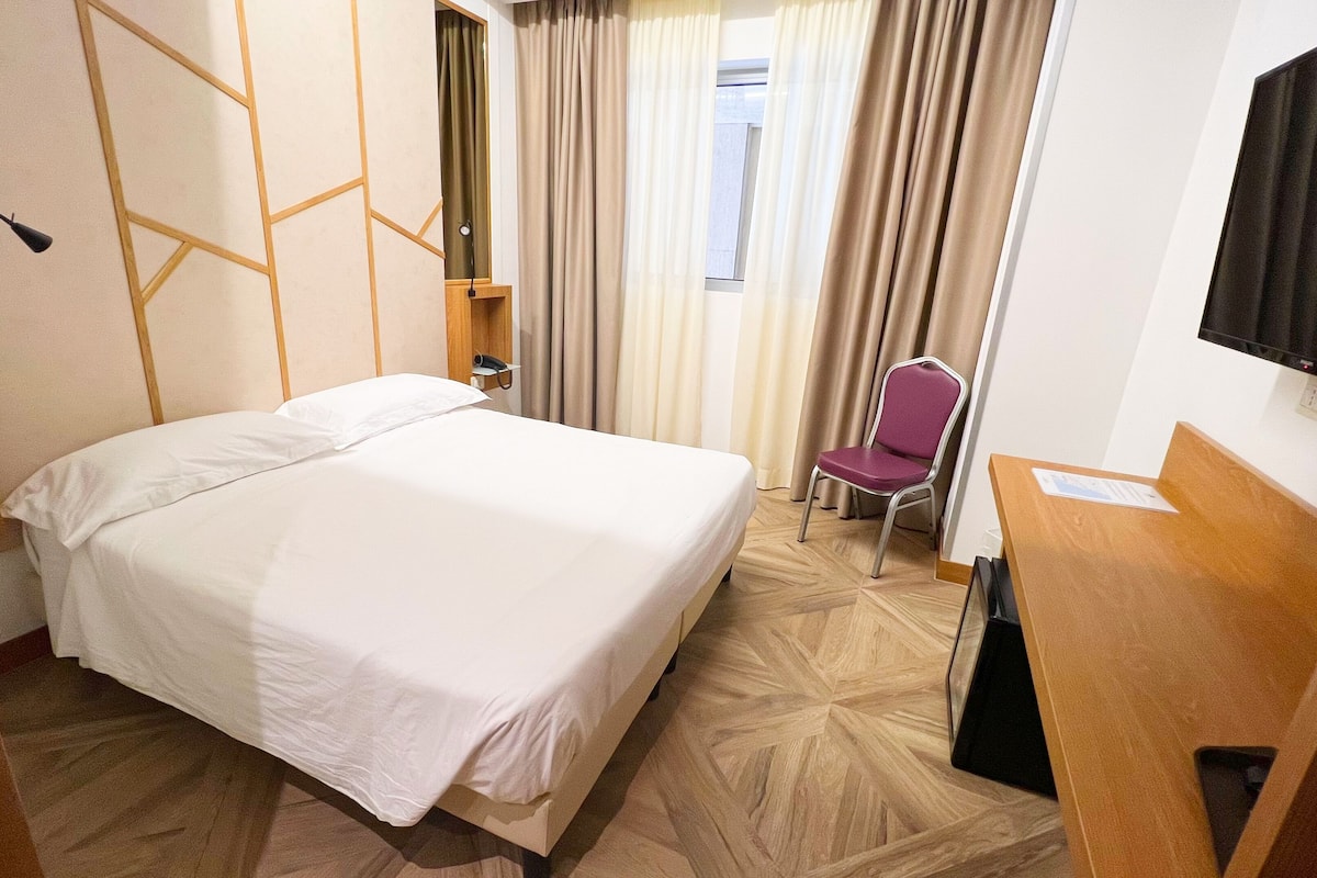 Een hotelkamer met een bed, een bureau, een stoel, een televisie en een raam met gordijnen.