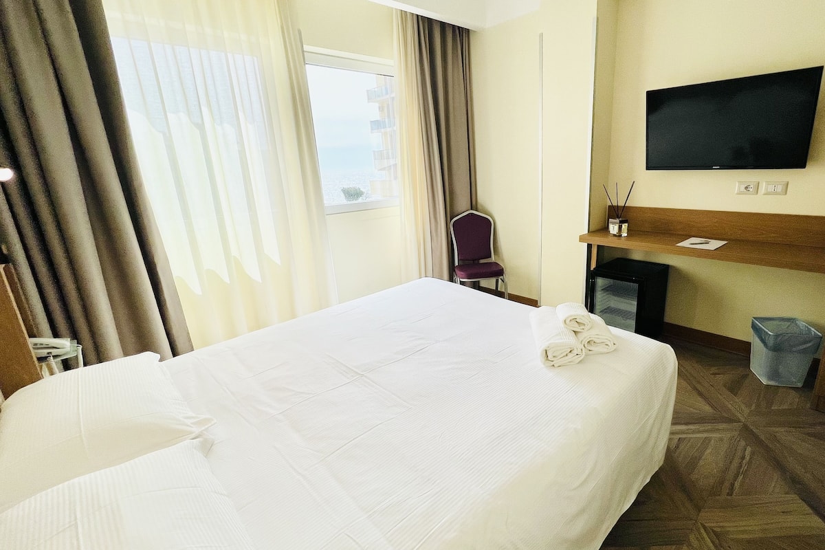 Een hotelkamer met een bed, een aan de muur bevestigde televisie, een stoel en een klein tafeltje met een lamp.