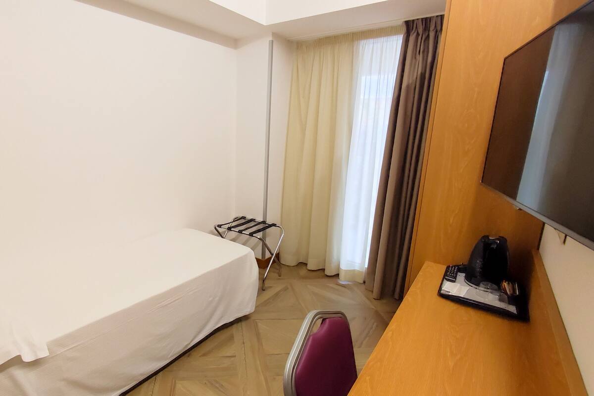 Een kleine hotelkamer met een eenpersoonsbed, een bureau met stoel, een televisie en een raam met gordijnen.