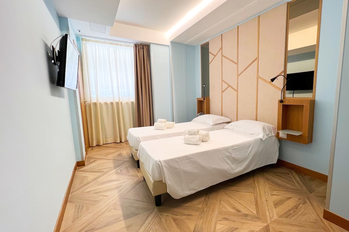 Een hotelkamer met twee bedden, een aan de muur bevestigde televisie, een houten vloer en een raam met gordijnen.