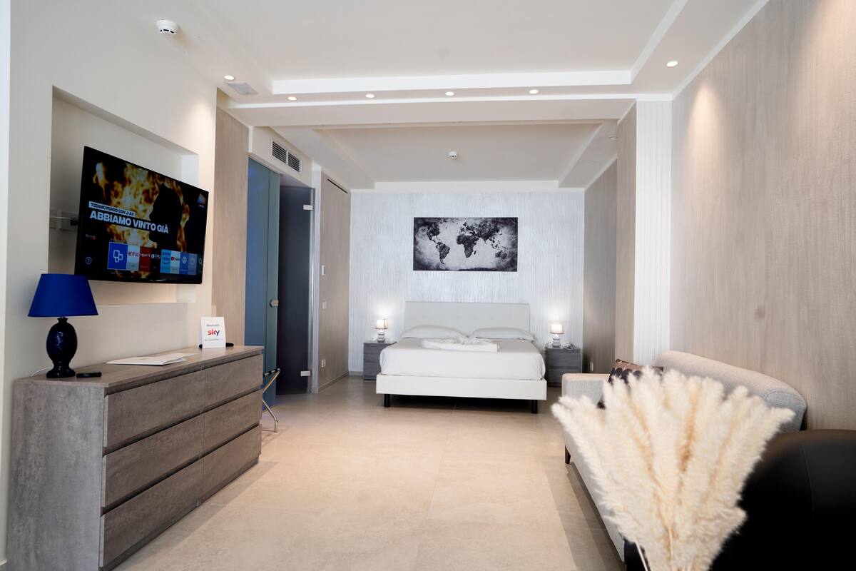Een moderne hotelkamer met een groot bed, een bureau met een flatscreen televisie, een nachtkastje met een lamp en een decoratieve plant.