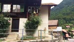 A two-story house with a stone wall in front, a metal railing, and a mountain backdrop. - Studio by Apartments Capricorn (Gsteigwiller)