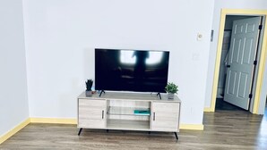A living room with a flat-screen TV on a wooden cabinet, a potted plant, and a door leading to another room. - Glacier Lake 2nd floor 2 bdrm condo (Joseph)
