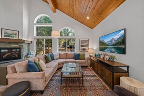 A spacious living room with a large sectional sofa, a glass coffee table, and a wooden cabinet. There are large windows providing natural light and views of the outdoors. - Chic Pet-Friendly Retreat | King Suite, Hot Tub | Clubhouse Access | Near Lake (Truckee)