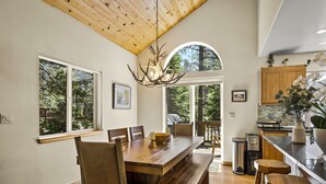 A spacious dining area with a wooden table and benches, large windows offering a view of the outdoors, and a kitchen area with wooden cabinets and a countertop. - Chic Pet-Friendly Retreat | King Suite, Hot Tub | Clubhouse Access | Near Lake (Truckee)