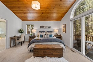 A bedroom with a large bed, wooden headboard, two bedside tables with lamps, a window, and a view of the outdoors.