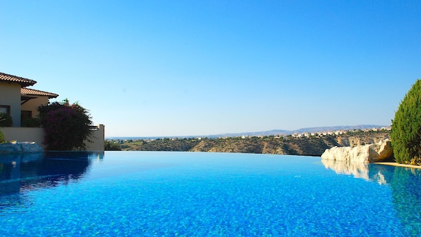 Une magnifique piscine à débordement d’un bleu profond, offrant une vue sur un paysage côtier et sur un bâtiment doté d’un toit recouvert de tuiles.