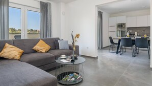 Ett modernt vardagsrum med en grå soffa, ett soffbord och en matplats, med ett kök synligt i bakgrunden.
