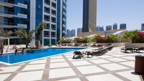 Un área de piscina en la azotea con sillones de descanso y palmeras, rodeada de edificios altos.