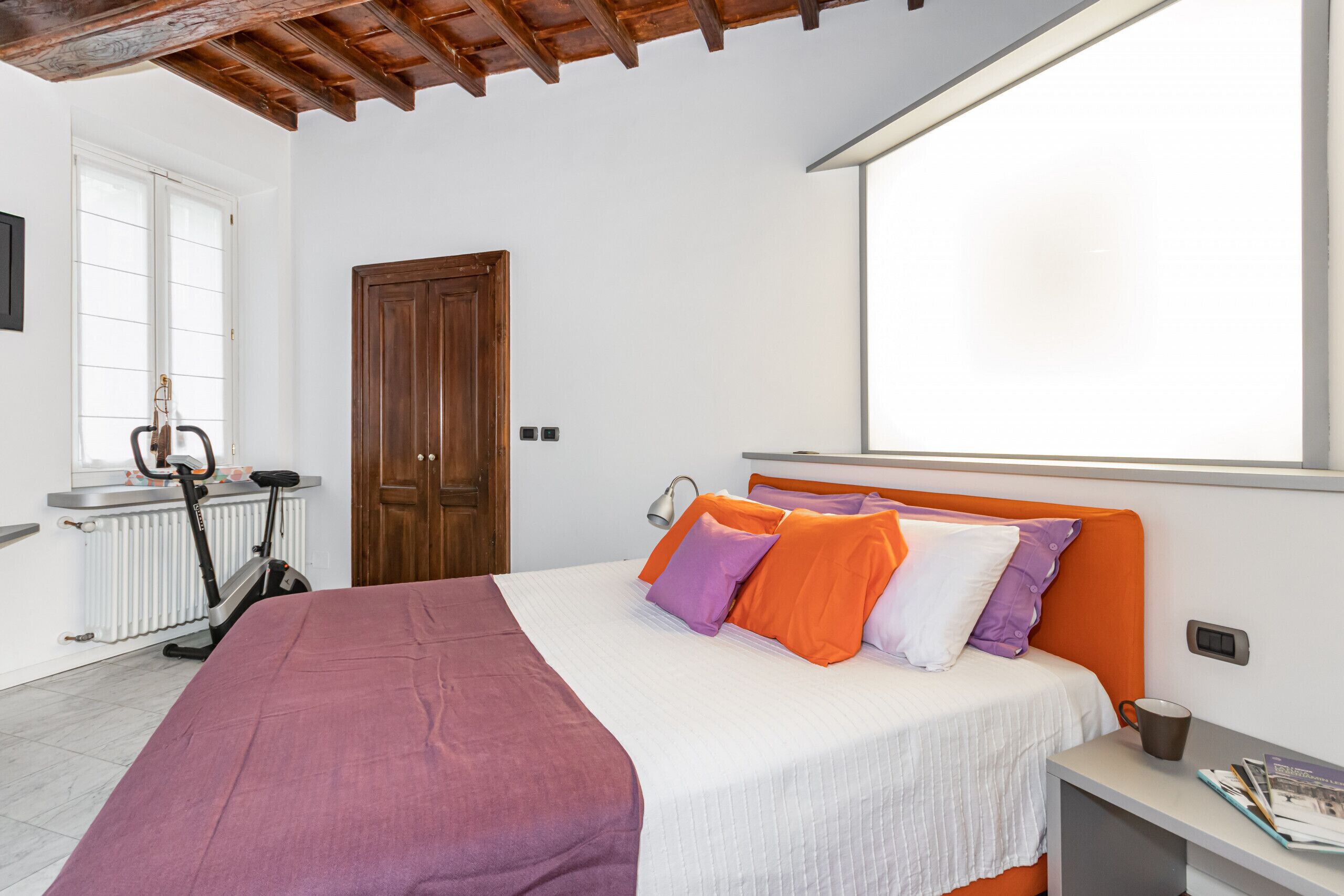 Ein Schlafzimmer mit einem Bett, einem Heimtrainer, einer Holztür und einem Fenster.
