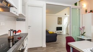 A modern kitchen with a stove, kettle, and fruit bowl, leading to a living area with a sofa, TV, and a dining table with chairs. - That's a Mole by Wonderful Italy (Torino)