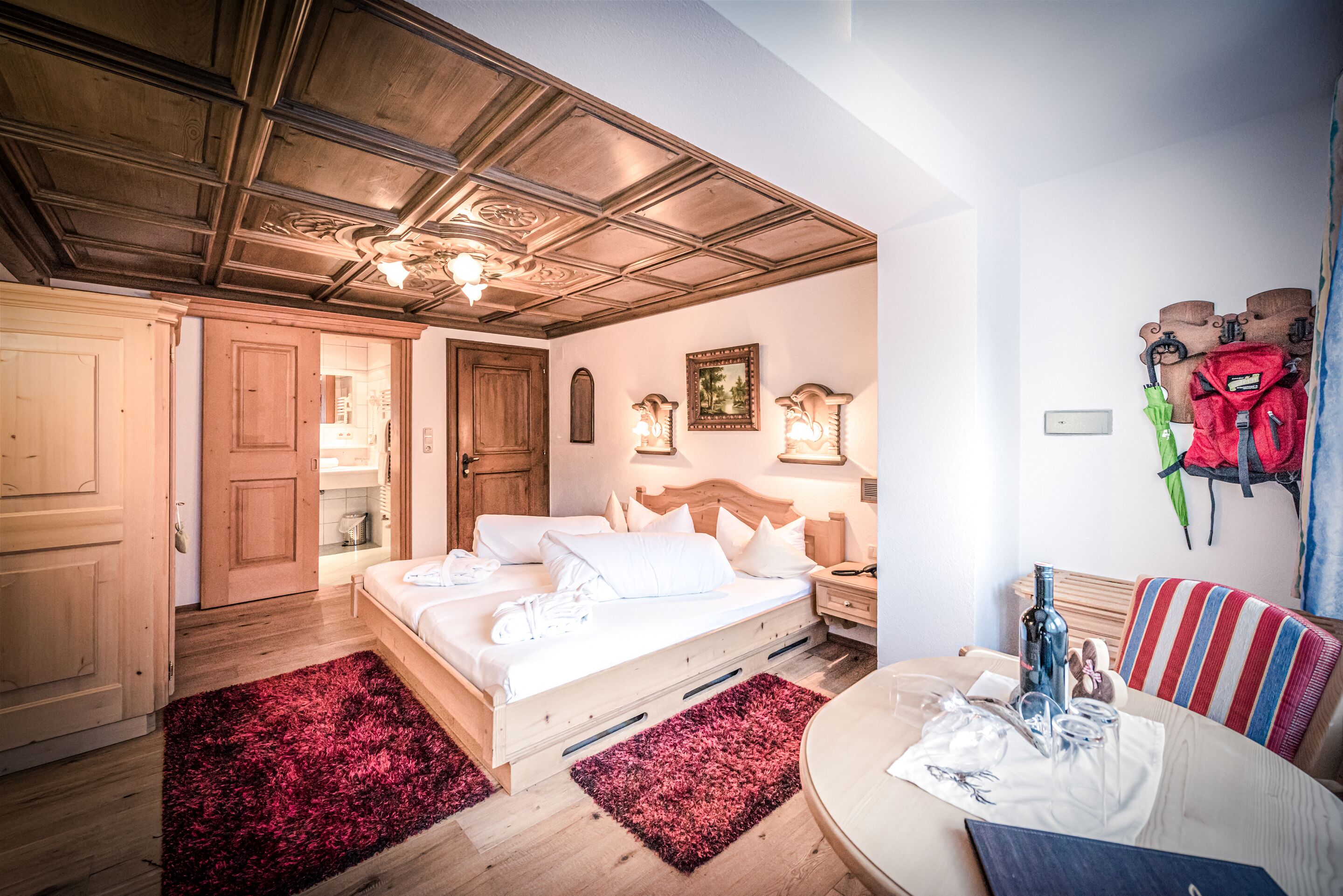 Un dormitorio con techo de madera, una cama con ropa de cama blanca, una alfombra roja, una mesita de noche con una botella y un perchero en la pared.