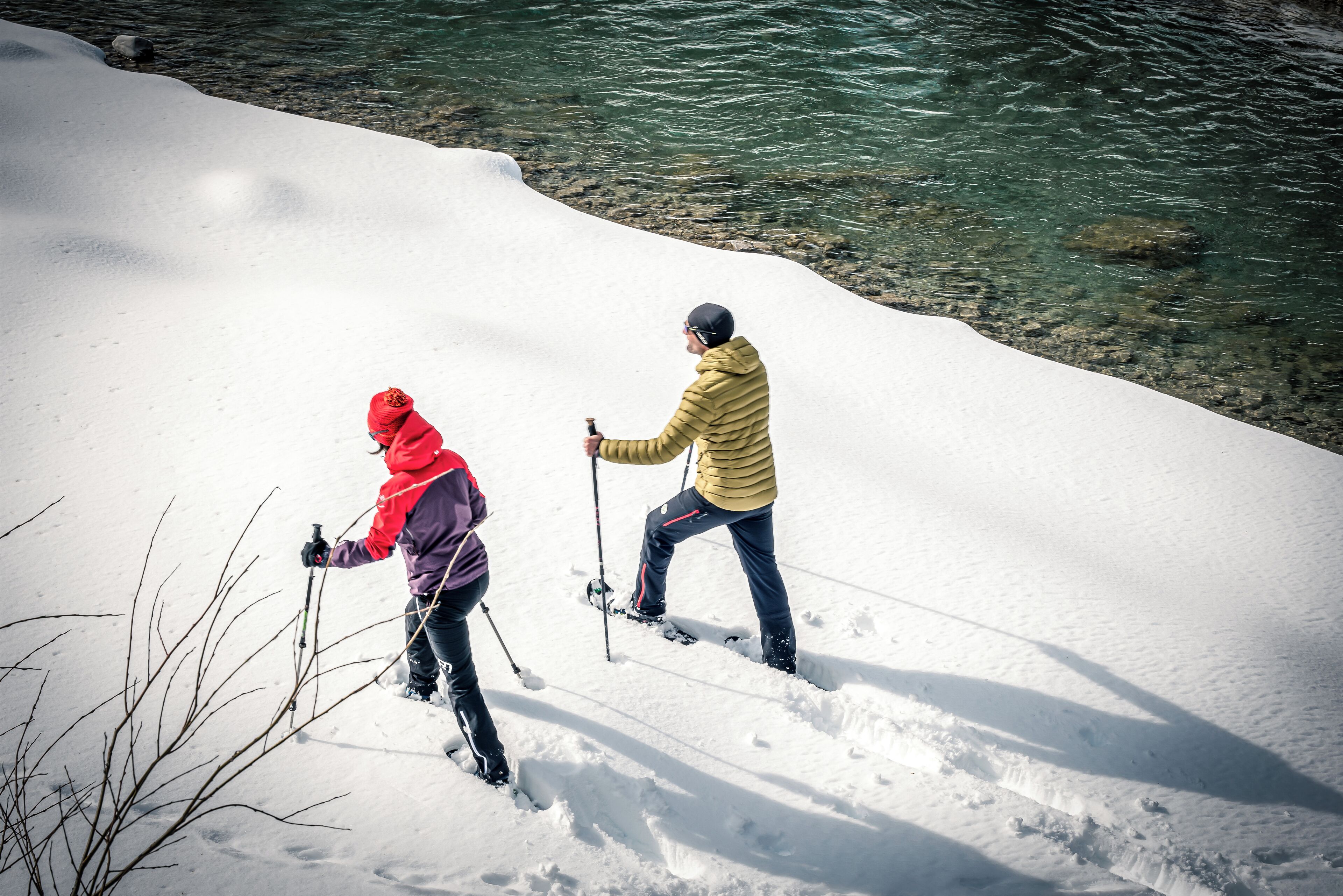 Two people cross-country skiing on a snowy slope with a clear water body in the background.