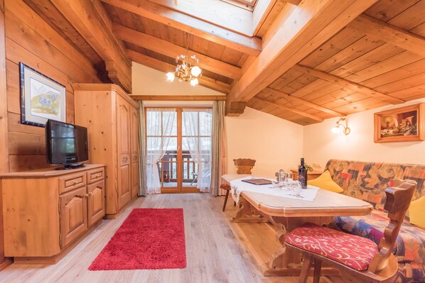 Interior de madera con una mesa de comedor, sillas, un tapete rojo, un televisor y una ventana con cortinas.