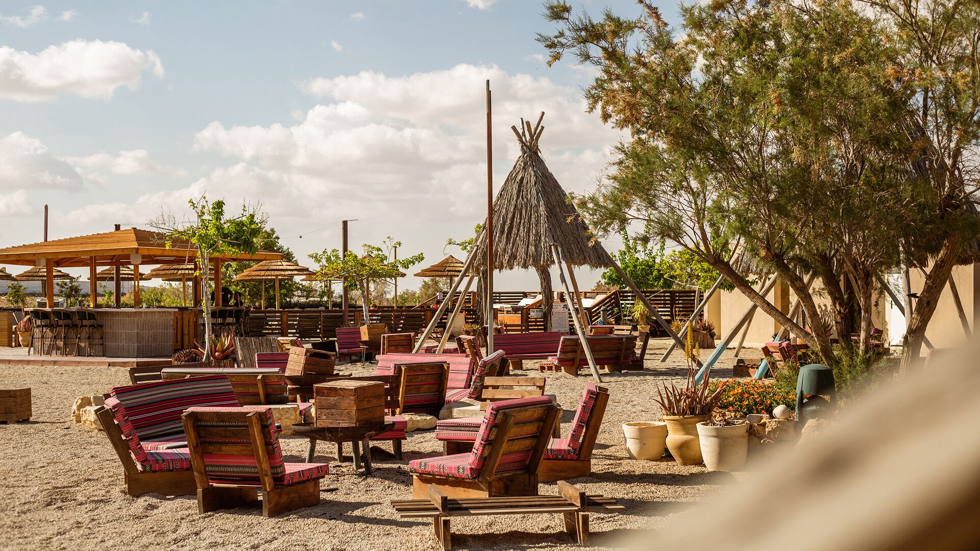 Outdoor seating with wooden chairs and tables, a thatched-roof structure, and a sandy ground.