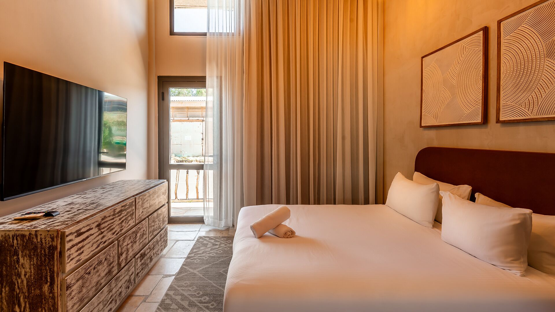 Habitación de hotel con una televisión de pantalla plana grande, un aparador de madera, una cama con ropa blanca y dos obras de arte colgadas en la pared.
