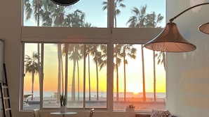 A spacious living room with large windows overlooking a beach at sunset, featuring a modern sofa, chairs, and a small table.
