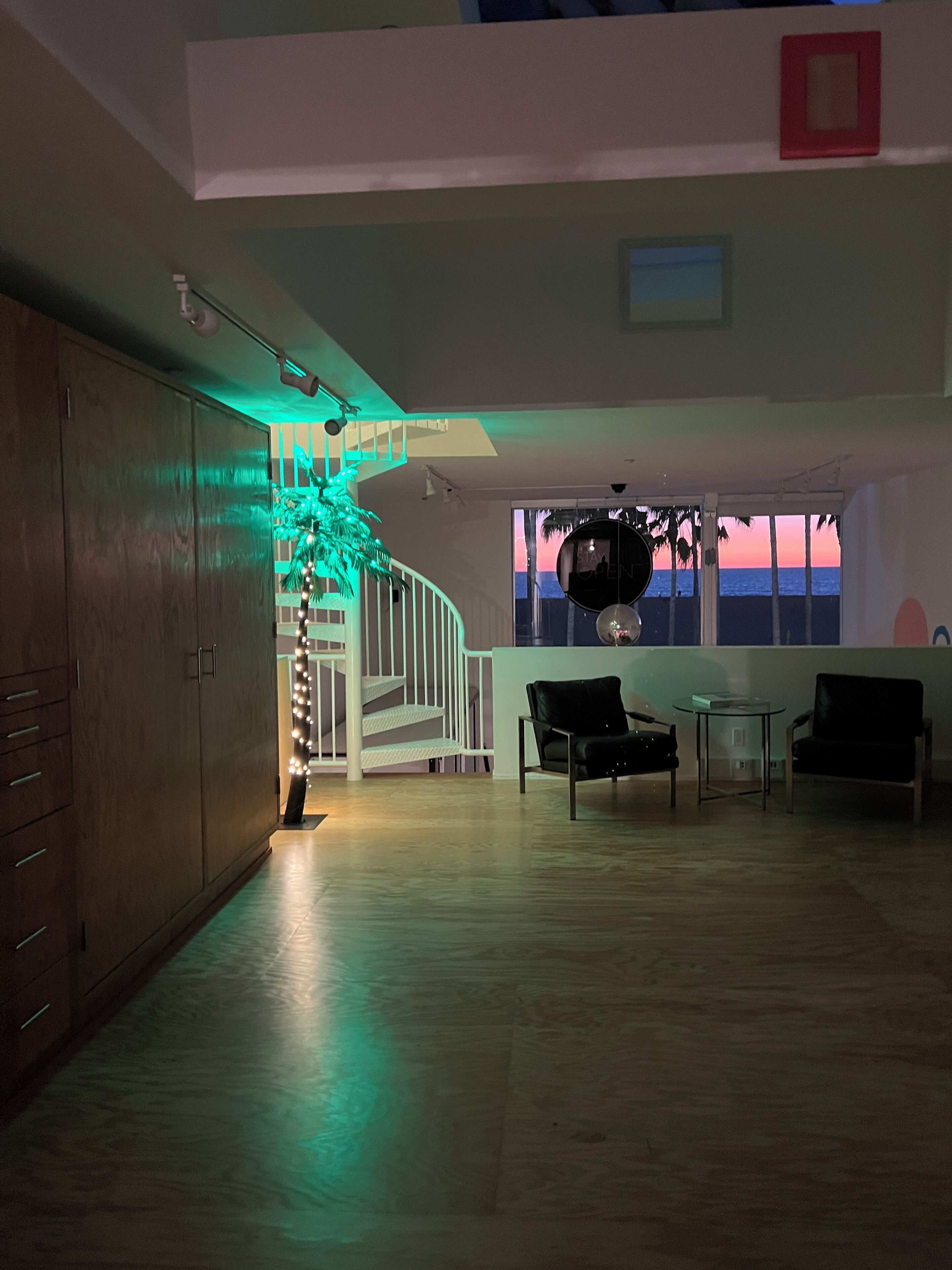 A spacious room with a staircase, a view of the sunset through a window, and a seating area with chairs and a table.